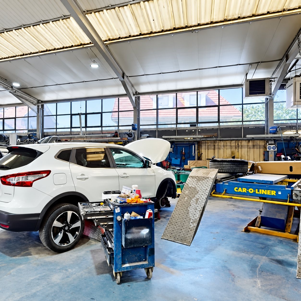 Intérieur d'un atelier de carrosserie où un SUV blanc au capot ouvert se trouve près d'un banc de redressage Car-O-Liner et d'un chariot d'outils bleu.