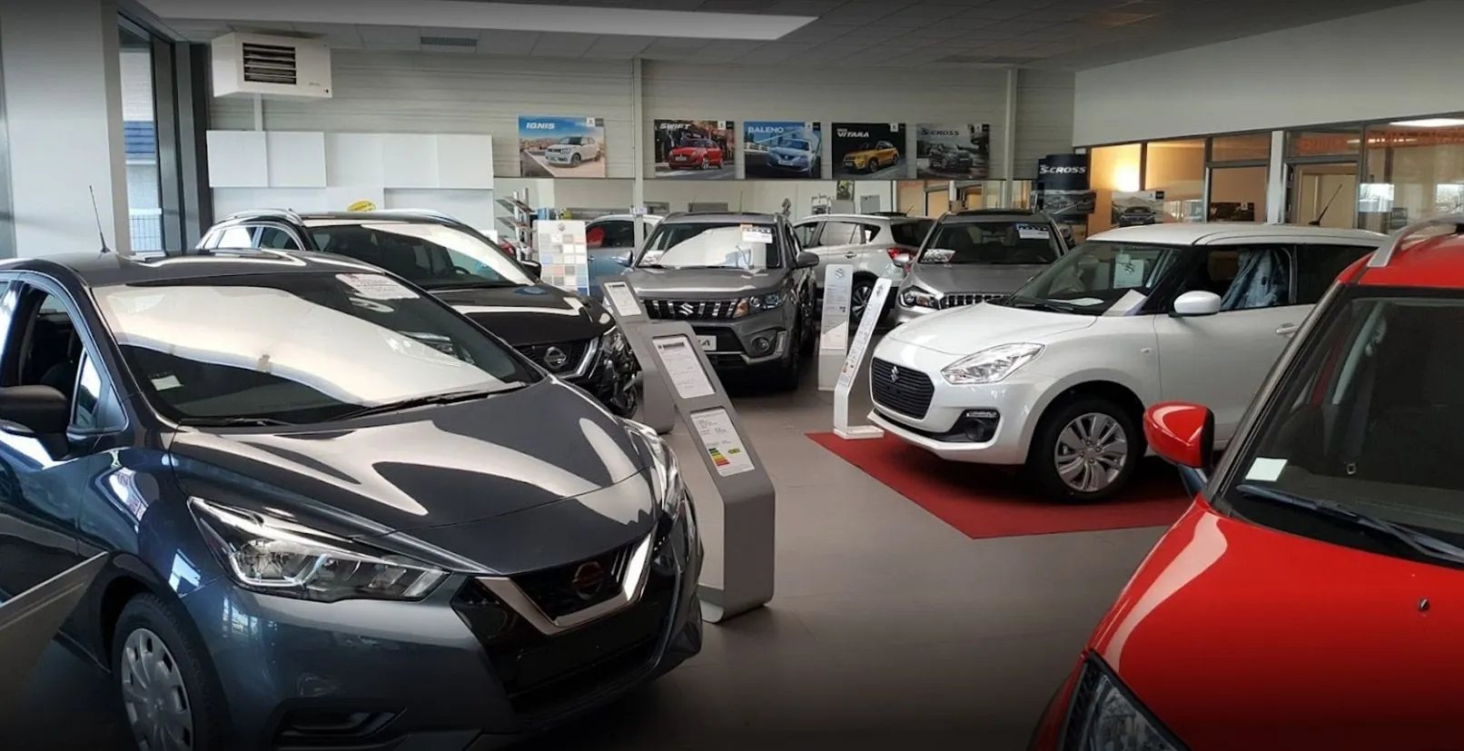 Intérieur lumineux d'un concessionnaire automobile où sont exposées des voitures grises, blanches et rouges, avec des affiches de modèles au mur.